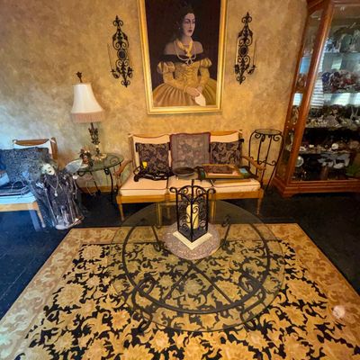 Sale Photo Thumbnail #3: Living room setup featuring a large framed portrait of a woman, wooden settee with patterned cushions, wrought-iron glass-top coffee table over a floral area rug, and a tall glass display cabinet.