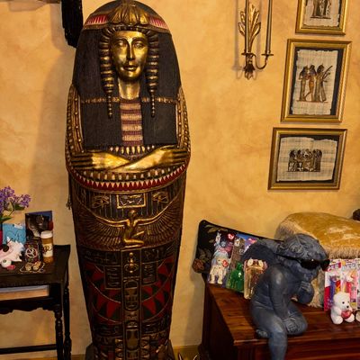 Sale Photo Thumbnail #111: Large Egyptian revival sarcophagus cabinet, painted in gold, red, and black, displayed alongside framed papyrus artwork and decorative home goods.