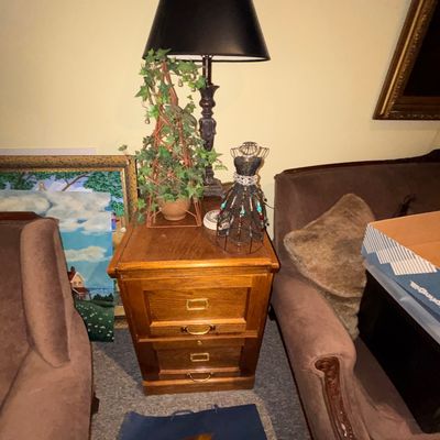 Sale Photo Thumbnail #91: Two-drawer oak file cabinet style end table topped with a lamp with a black shade, a faux ivy topiary, and a decorative mini wire dress form, flanked by brown velvet armchairs.