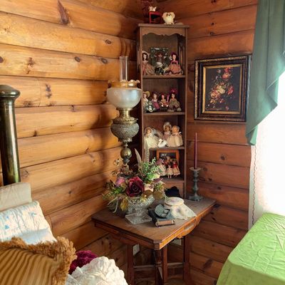 Sale Photo Thumbnail #74: Vintage log cabin bedroom corner featuring a wooden drop-leaf table, an ornate brass oil-style electric lamp, and a tall narrow bookshelf filled with collectible dolls.