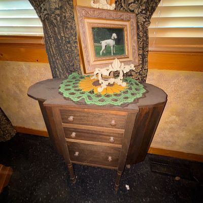 Sale Photo Thumbnail #31: Vintage drop-leaf wooden sewing cabinet or side table with three drawers, topped with a colorful crochet doily and framed dog portrait.