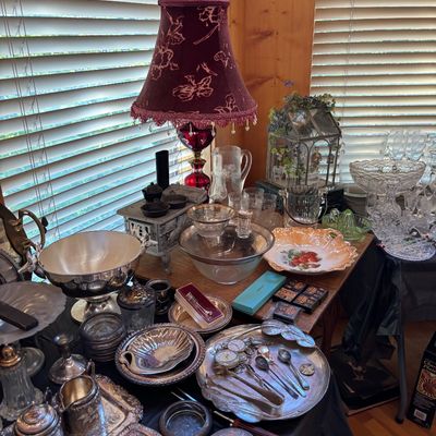 Sale Photo Thumbnail #69: A collection of vintage silver-plated serving pieces, glassware, and decorative tableware displayed on a table alongside a red fabric-shaded lamp.