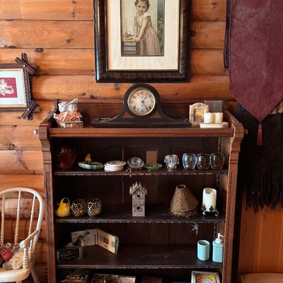 Sale Photo Thumbnail #63: Vintage dark wood bookshelf filled with small collectibles, trinkets, and glassware, set against a log cabin wall with framed art and a mantle clock above.