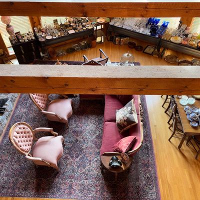 Sale Photo Thumbnail #76: High-angle view of a living area featuring vintage tufted armchairs, a red sofa, a dining table set with china, and perimeter tables filled with glassware and collectibles.