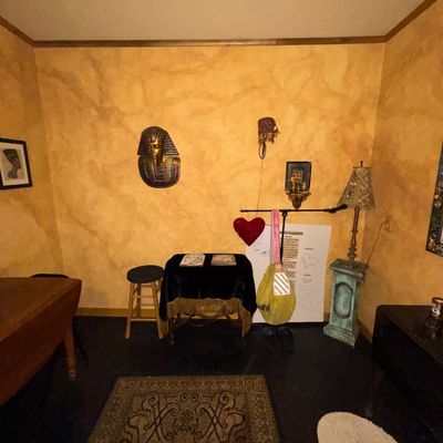 Sale Photo Thumbnail #118: Eclectic interior setup featuring an Egyptian King Tut wall bust, vintage table lamp on a pedestal, wooden furniture, and patterned area rugs.