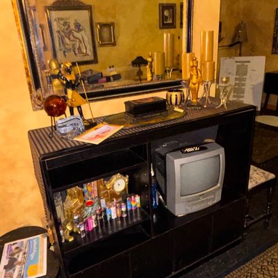 Sale Photo Thumbnail #95: Black wooden shelving unit featuring Egyptian-themed decor, a small CRT television combo, collection of lighters, and a large gilt-framed wall mirror hung above.