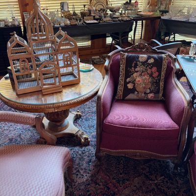 Sale Photo Thumbnail #78: Traditional burgundy upholstered arm chair with carved wood trim paired with a decorative architectural wooden birdcage on a round pedestal table.