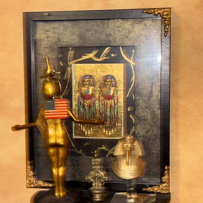 Sale Photo Thumbnail #113: Egyptian-revival decor display featuring a framed art piece with beaded pharaoh accents, a gold-tone statuette of Isis wearing a small American flag pin, and a mini King Tut bust on a black wall shelf.
