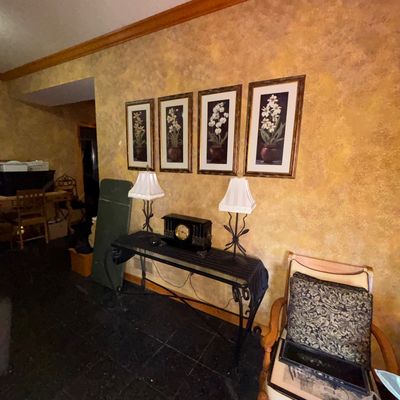 Sale Photo Thumbnail #86: Wrought iron console table featuring a dark wooden mantel clock, flanked by matching table lamps, with a set of four framed floral prints mounted on the wall above.