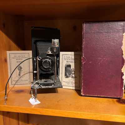 Sale Photo Thumbnail #23: Vintage Seneca folding bellows camera displayed with original instructional booklets and a burgundy storage box on a wooden shelf.