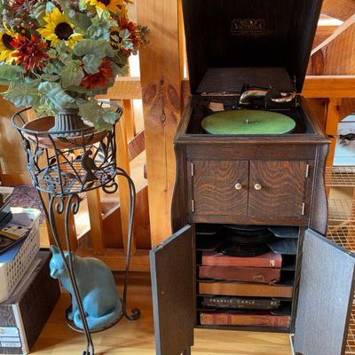 Sale Photo Thumbnail #50: Antique Victrola oak floor-standing phonograph cabinet with open doors showing record storage, paired with a wrought iron plant stand holding floral decor.