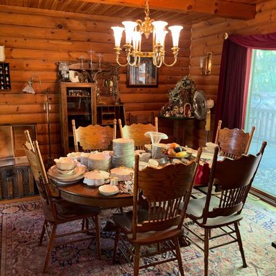 Sale Photo Thumbnail #56: Traditional oak dining table and press-back chairs set, featuring a large collection of china, situated in a log cabin room under a brass chandelier.