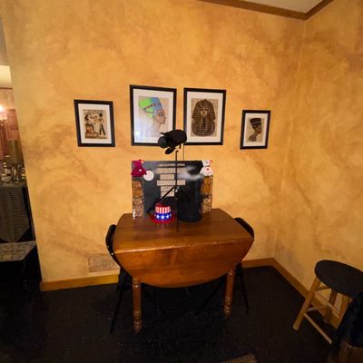Sale Photo Thumbnail #116: Small wooden drop-leaf table paired with folding chairs, set against framed Egyptian-themed wall art and featuring novelty items atop the table.