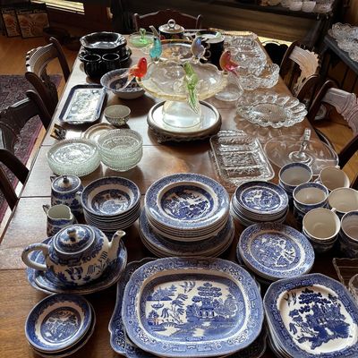 Sale Photo Thumbnail #70: Extensive collection of vintage Blue Willow pattern china tableware displayed alongside clear press glassware and colorful art glass pieces on a wooden dining table.