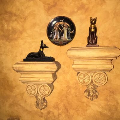 Sale Photo Thumbnail #96: Egyptian-revival wall display featuring decorative stone-look corbels supporting black and gold Anubis and Bastet figurines, alongside a decorative round wall plate.