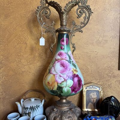 Sale Photo Thumbnail #14: Ornate antique hand-painted floral porcelain vase mounted with elaborate brass double handles and pedestal base. Displayed alongside a ceramic tea set, framed portrait, and small decor items.