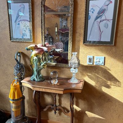 Sale Photo Thumbnail #72: French Provincial style console table with a large colorful parrot figurine, glass table lamp, arched ornate wall mirror, pair of framed bird prints, and a yellow Dyson upright vacuum cleaner.