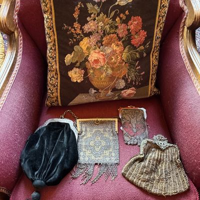 Sale Photo Thumbnail #127: Collection of vintage and antique purses, including beaded mesh and black velvet styles, displayed on an armchair alongside a floral tapestry throw pillow.