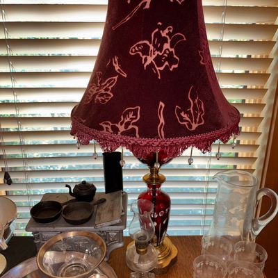Sale Photo Thumbnail #8: Vintage ruby flash cut glass table lamp featuring a burgundy velvet floral shade with fringe detailing, surrounded by assorted glassware and mini cast iron cookware.