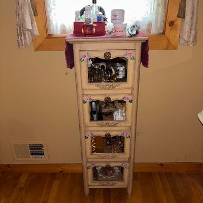 Sale Photo Thumbnail #43: Vintage painted wooden storage tower featuring four glass-panel door compartments with hand-painted floral accents and brass-tone hardware, housing various vanity items and perfume bottles.