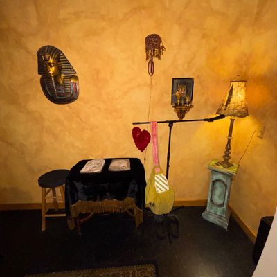 Sale Photo Thumbnail #114: Eclectic room decor including an Egyptian King Tut wall bust, a table with fringe cover, a tall floor lamp on a pedestal, and various hanging wall items.