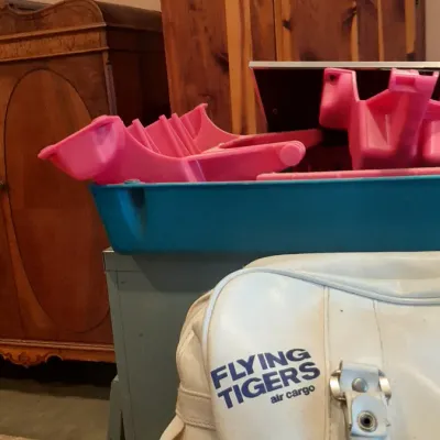 Vintage white Flying Tigers air cargo vinyl flight bag displayed in front of a blue storage tub with pink plastic components and wooden...