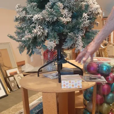 Pre-lit flocked tabletop artificial Christmas tree displayed on a particle board side table alongside a tall container of colorful...