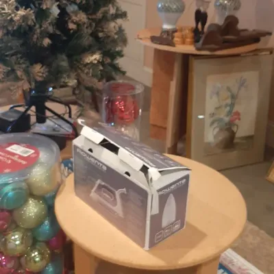 Boxed Rowenta steam iron resting on a light wood round side table, surrounded by a frosted artificial Christmas tree, colorful ornaments,...
