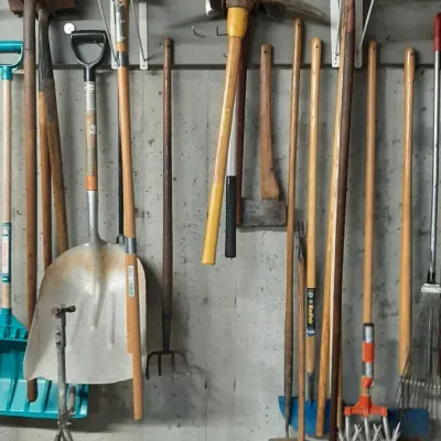 Collection of long-handled yard and garden tools, including shovels, rakes, a pickaxe, axes, and a green broadcast spreader.