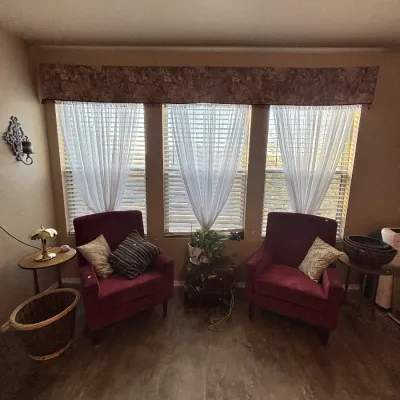 Sale Photo Thumbnail #20: Pair of burgundy velvet upholstered armchairs with throw pillows, flanked by small side tables beneath a window setup with a valance.