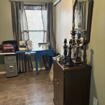 Sale Photo Thumbnail #25: Bedroom featuring a dark wood cabinet topped with candlestick lamps and a wall mirror, alongside a blue vanity table with sewing machines near the window.