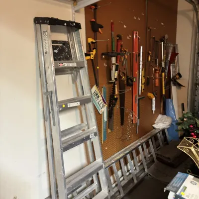 Sale Photo Thumbnail #3: An assortment of workshop tools, including aluminum step and extension ladders, clamps, handsaws, levels, and various hand tools organized on a pegboard.