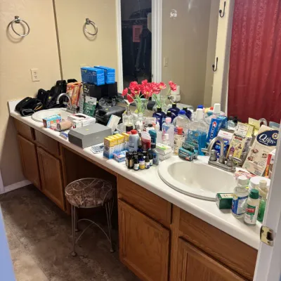 Sale Photo Thumbnail #16: A double-sink bathroom vanity filled with a variety of personal care products, toiletries, and health supplies, along with a small metal vanity stool.