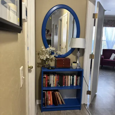 Sale Photo Thumbnail #27: Blue-painted small two-shelf bookcase filled with books, topped with a matching blue oval wall mirror, table lamp, and decorative accents.