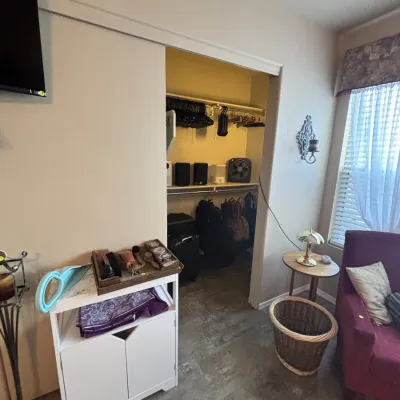 Sale Photo Thumbnail #19: Bedroom corner featuring a white accent cabinet topped with shoeshine supplies, a purple armchair, a small side table with a brass lamp, and an open closet filled with luggage, clothing, and small electronics.