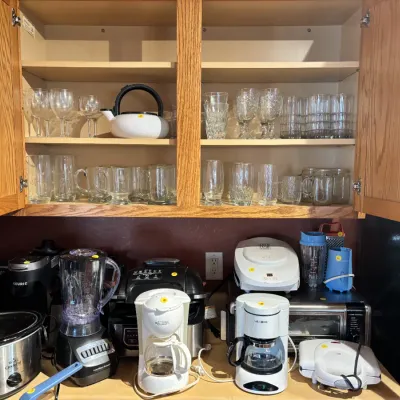 Sale Photo Thumbnail #11: Collection of small kitchen counter appliances and glassware arranged inside and beneath wooden cabinets, including coffee makers, blenders, a slow cooker, and various drinkware.