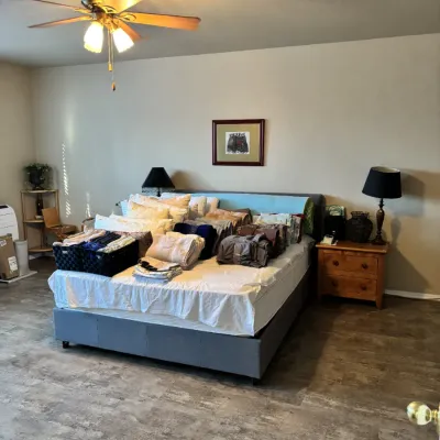 Sale Photo Thumbnail #22: Upholstered platform bed displayed with various linens, towels, and decorative pillows alongside a wooden nightstand with a dark-shaded table lamp.