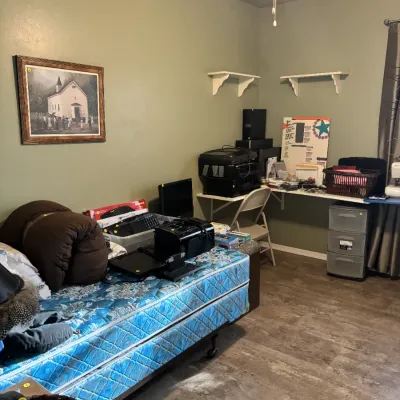 Sale Photo Thumbnail #26: Bedroom set up for sale featuring a blue patterned mattress loaded with office items, a wall-mounted desk space, and a framed church painting.