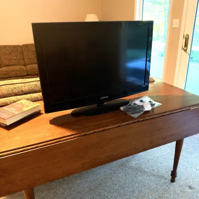 Black Samsung flat-screen television resting on a mid-century wooden drop-leaf table, with power cables in a plastic bag nearby.