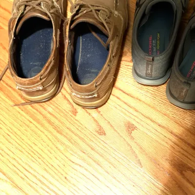 Two pairs of men's casual shoes resting on hardwood flooring, including tan leather boat shoes and dark grey Skechers sneakers.