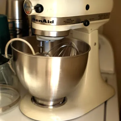 Off-white KitchenAid tilt-head stand mixer with a stainless steel mixing bowl and wire whisk attachment.