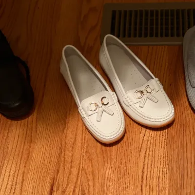 Pair of women's white leather loafers featuring brass bit hardware and tassel details, resting on a hardwood floor.