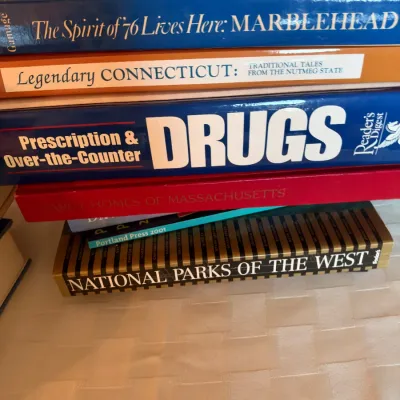 Stack of assorted hardcover coffee table and reference books, including titles on New England, prescription drugs, and National Parks.