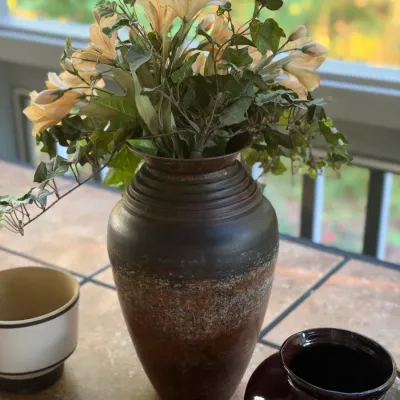 Large dark ceramic urn-style vase filled with faux flowers, displayed on a tiled surface alongside assorted small pottery pieces.