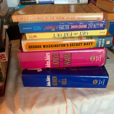 Stack of assorted hardcover and paperback books, including historical fiction by John Jakes, cookbooks, and military history titles.