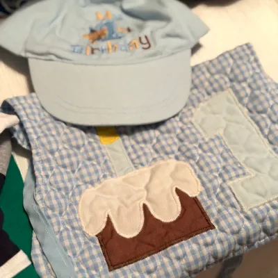 Baby boy's 1st birthday theme set featuring a light blue embroidered hat and matching blue gingham quilted bib with a cake appliqué.