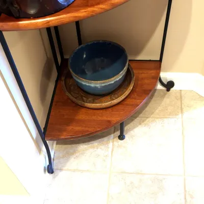 Blue glazed ceramic bowl sitting on a decorative brass tray atop the lower shelf of a corner metal and wood plant stand.