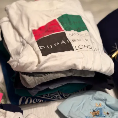 Folded stack of graphic and plain t-shirts, topped with a white graphic tee, alongside a light blue embroidered baseball cap.