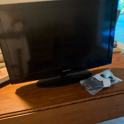 Black Samsung flat-screen LCD television resting on a wooden console, accompanied by a bagged power cord and accessories.