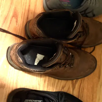 Pair of worn brown leather hiking boots resting on a hardwood floor alongside other casual athletic shoes.